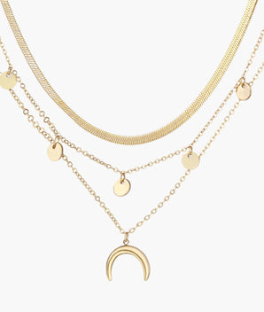 Four-layer gold necklace with herringbone chain, disc charms, and crescent moon pendant on cream background