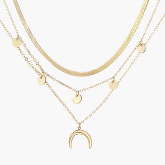 Four-layer gold necklace with herringbone chain, disc charms, and crescent moon pendant on cream background