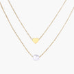 Two delicate gold-plated chains with heart charm and white pearl bead on cream background, minimal layered design.