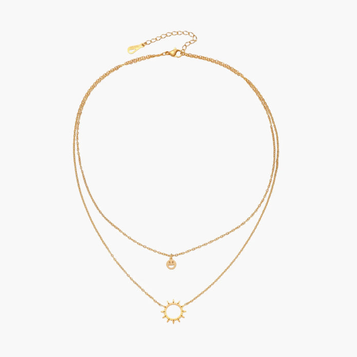 Gold layered necklace set with sunburst and smiley face pendants on delicate chains against white background.