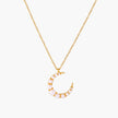 Gold crescent moon pendant with graduated white pearls on delicate chain against cream background