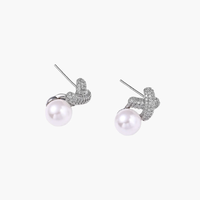 White gold plated studs featuring pavé X-shaped tops with freshwater pearl drops on cream background.