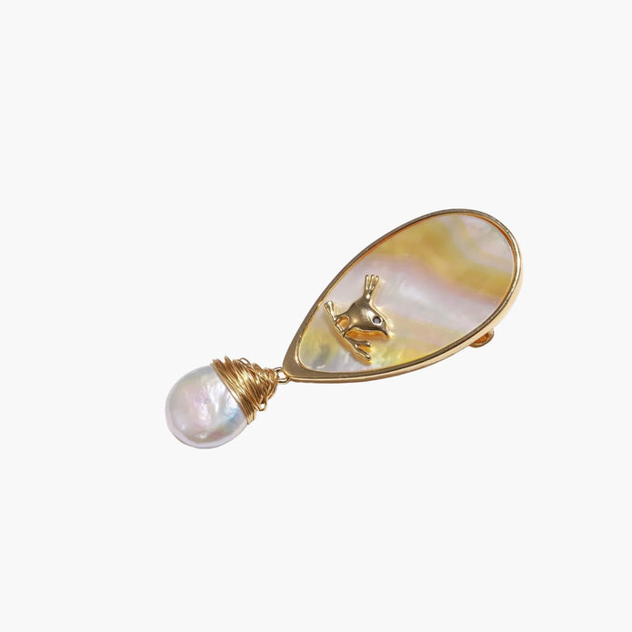 Iridescent shell brooch with gold bird accent and wire-wrapped baroque pearl drop on cream background.