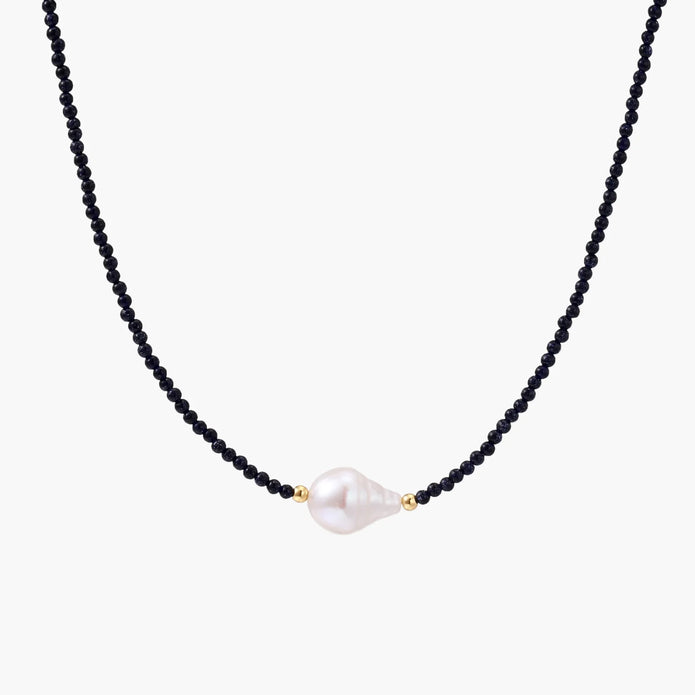 Delicate necklace with black blue sandstone beads and baroque pearl center on white background with gold accents.