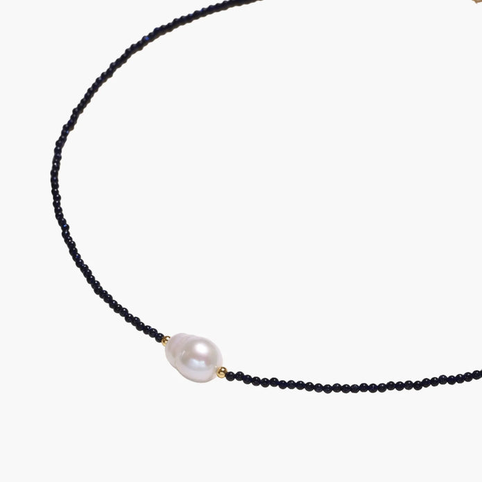 Delicate necklace with black blue sandstone beads and baroque pearl center on white background with gold accents.