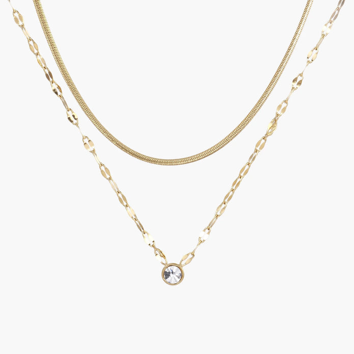 Layered gold necklace with herringbone and link chains, small bezel-set crystal pendant on cream background