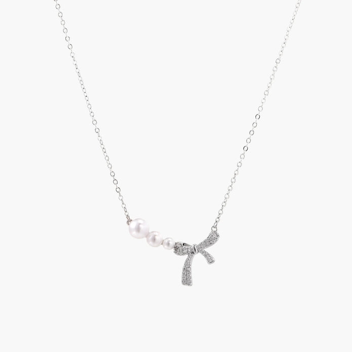 Delicate white gold plated necklace with graduated freshwater pearls and crystal-studded bow pendant on ivory background