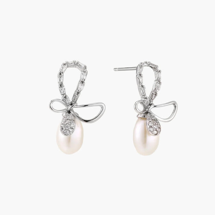 Silver bow-shaped stud earrings with baguette crystals and dangling white freshwater pearls on cream background