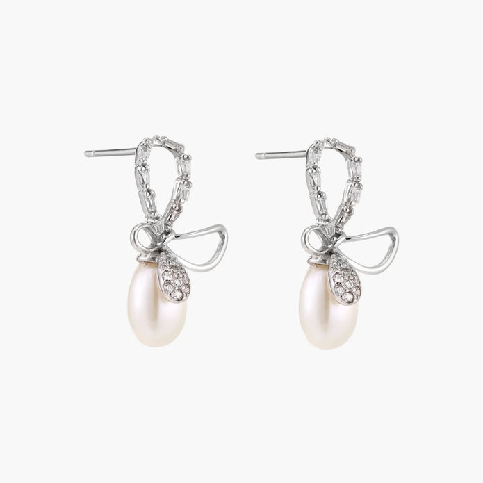 Silver bow-shaped stud earrings with baguette crystals and dangling white freshwater pearls on cream background