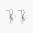 Silver bow-shaped stud earrings with baguette crystals and dangling white freshwater pearls on cream background
