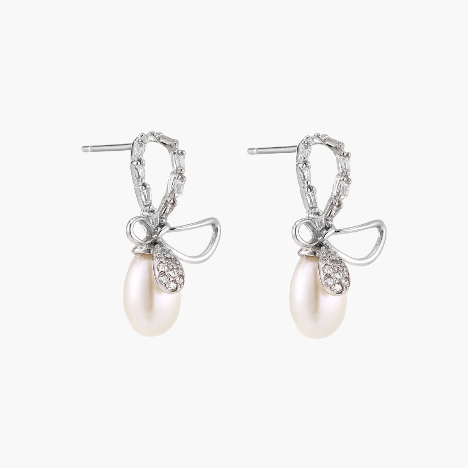 Silver bow-shaped stud earrings with baguette crystals and dangling white freshwater pearls on cream background