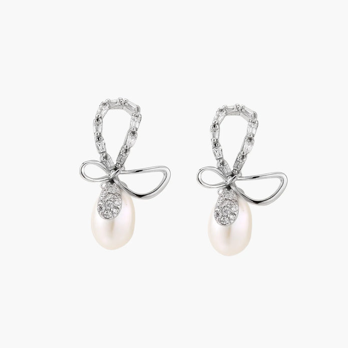 Silver bow-shaped stud earrings with baguette crystals and dangling white freshwater pearls on cream background