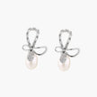 Silver bow-shaped stud earrings with baguette crystals and dangling white freshwater pearls on cream background