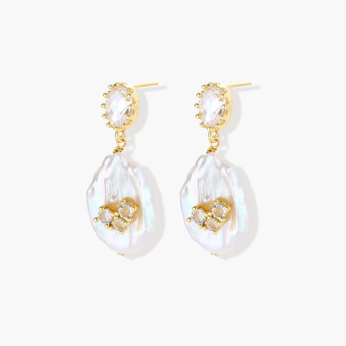 Gold-plated drop earrings with iridescent baroque pearls and crystal flower accents on cream background