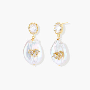 Gold-plated drop earrings with iridescent baroque pearls and crystal flower accents on cream background