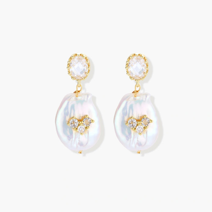 Gold-plated drop earrings with iridescent baroque pearls and crystal flower accents on cream background