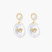 Gold-plated drop earrings with iridescent baroque pearls and crystal flower accents on cream background