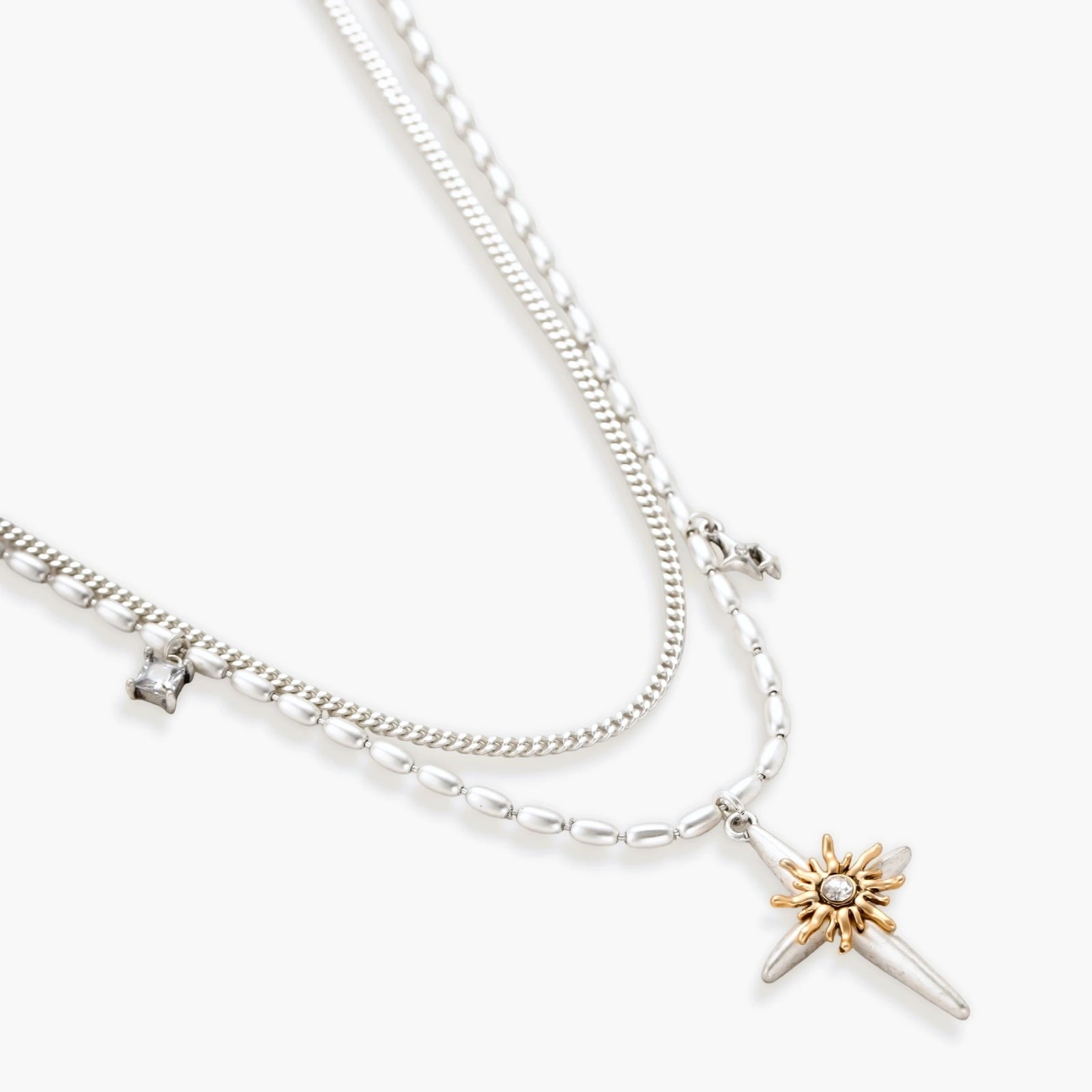 Layered silver-toned necklace set with cross pendant and gold sun charm detail, matching drop earrings on white background.