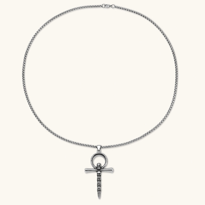 Silver stainless steel ankh pendant with skull motifs on vertical bar and textured ring top, on a box chain necklace.
